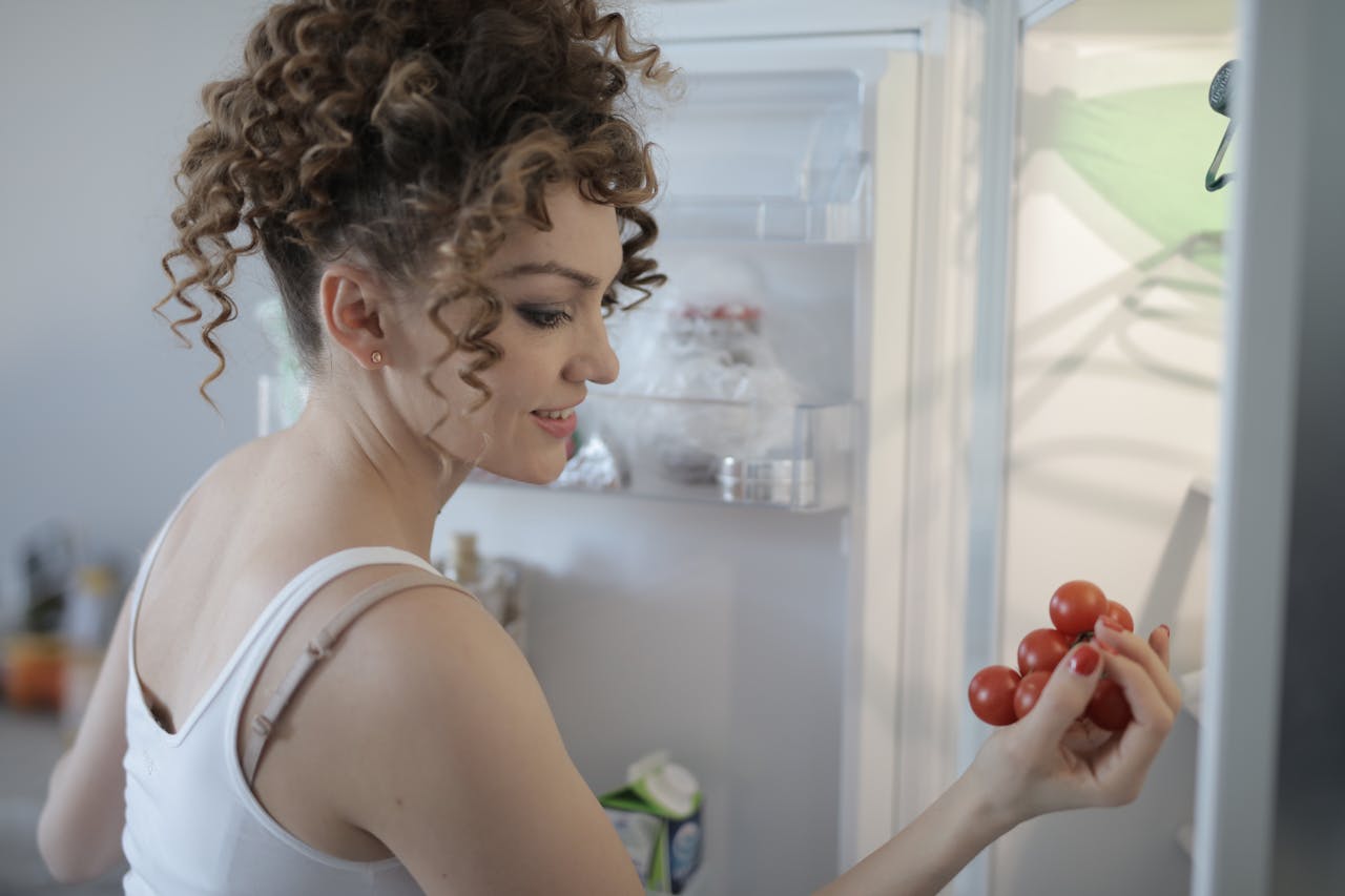 Side view of content female in casual wear standing in kitchen near opened fridge and picking fruit for breakfast